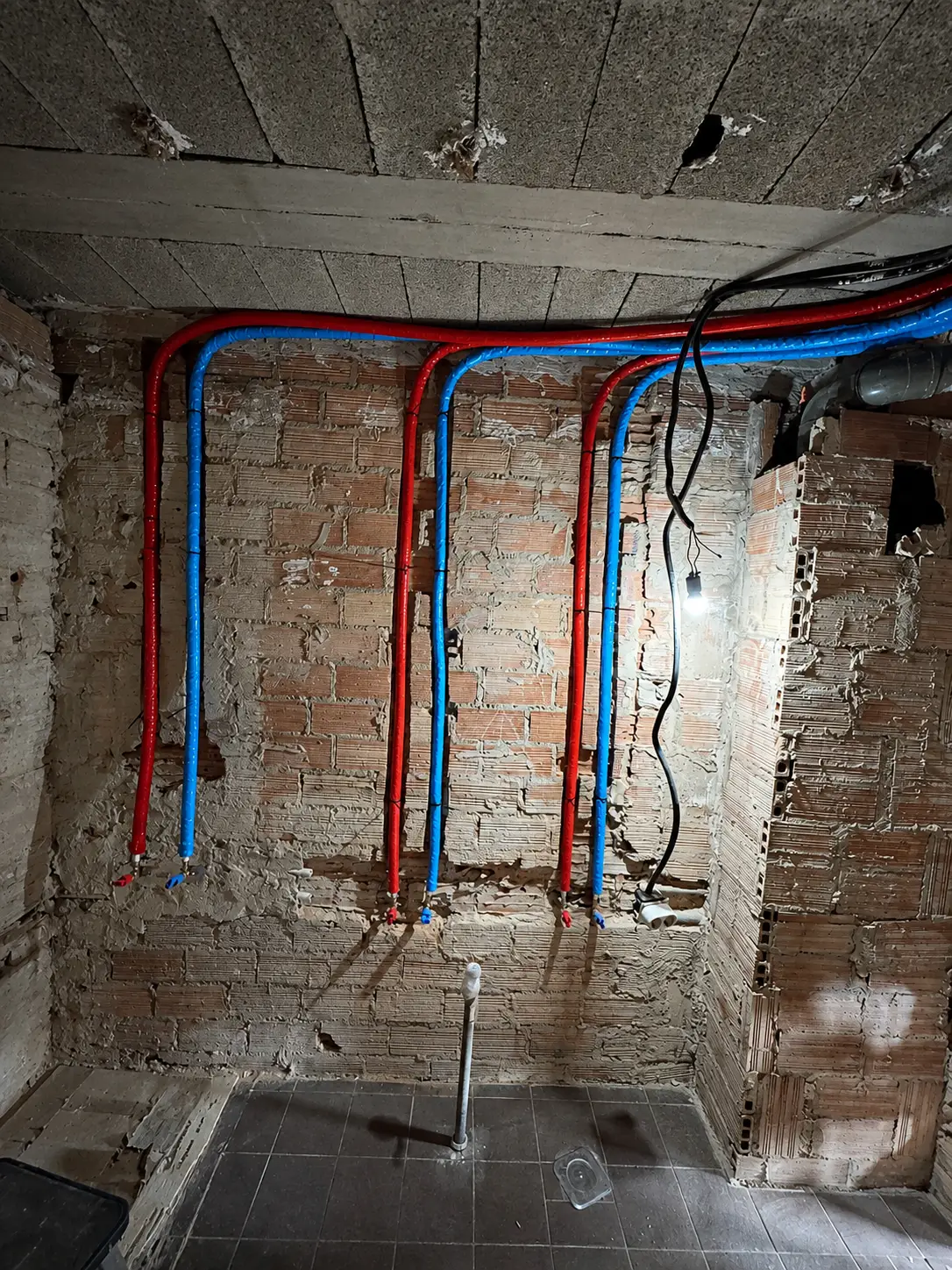 New red and blue water supply pipes installed on an exposed brick wall during bathroom renovation in Valencia.